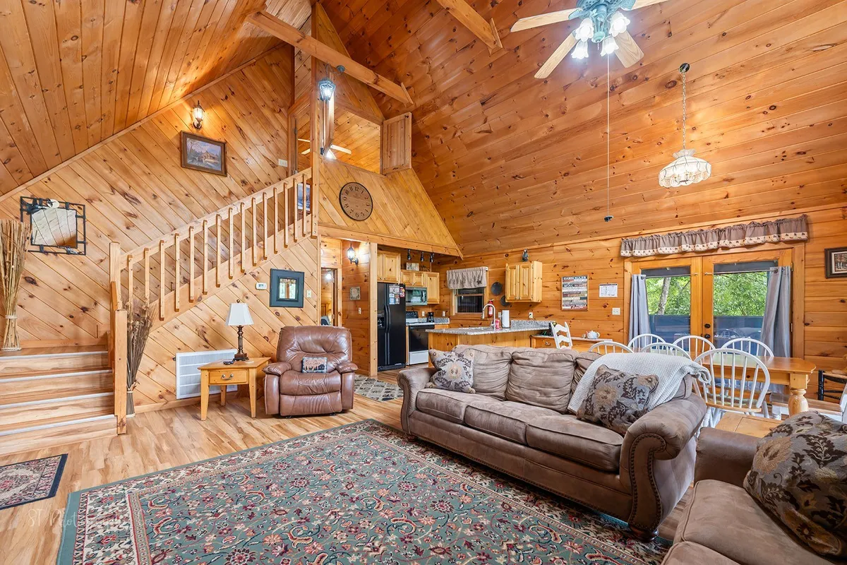 This image features a seamless open-concept space that includes the living room seating, dining area, kitchen, and a wooden staircase, all illuminated by natural light. The room is anchored by a cozy fireplace and arranged to encourage group gatherings and comfortable lounging.
