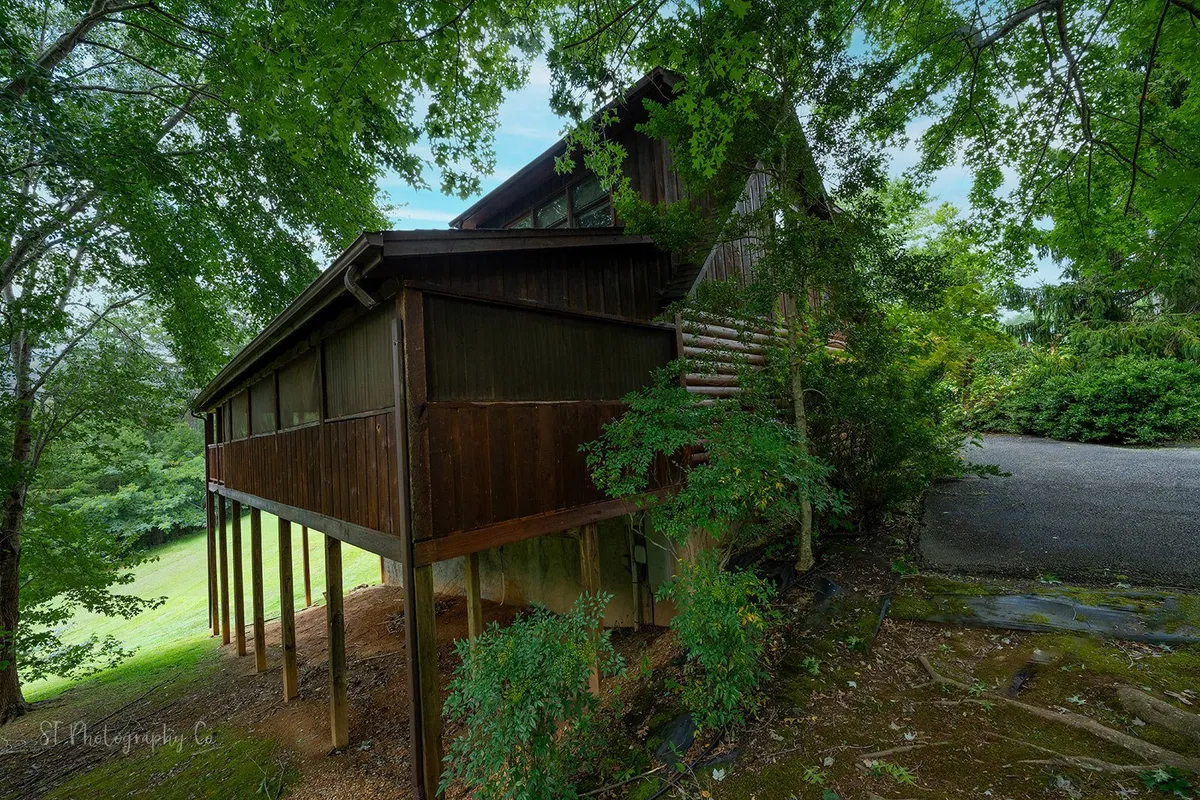 A striking wooden cabin elevated on sturdy stilts, nestled among lush greenery with a sloped mountain landscape in the background. The spacious porch offers unobstructed views of the natural surroundings, where wildlife can sometimes be spotted roaming the yard. The changing seasons are visible in the foliage and sunset colors.