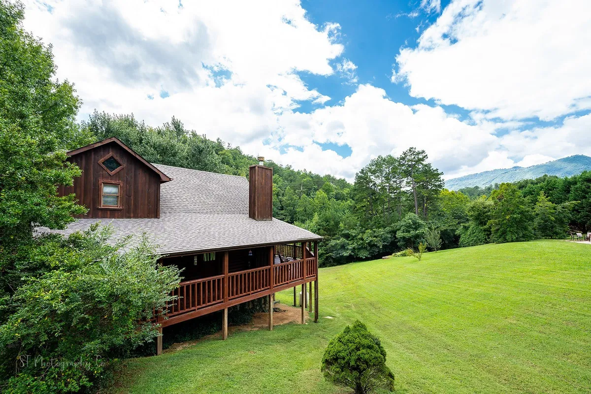 A wide exterior view showcasing the wooden house surrounded by an expansive green lawn, tall trees, and distant mountain scenery. The porch and open sky hint at opportunities for outdoor relaxation and exploration.