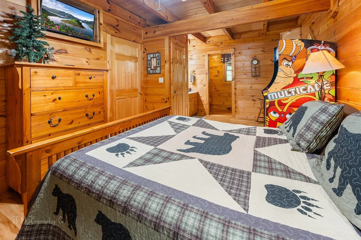 This playful master suite features a bed with a bear-themed quilt, a wooden dresser, a full-size arcade game machine, and framed landscape art. The space feels cozy yet vibrant, perfect for both relaxation and entertainment.