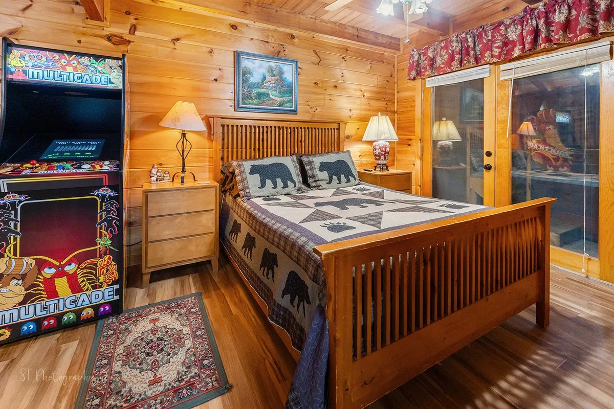 A fun, family-friendly bedroom with a bed featuring bear-themed bedding, wooden nightstands, and a sliding glass door providing deck access and scenic views. A multicade arcade machine adds a playful touch to the space.