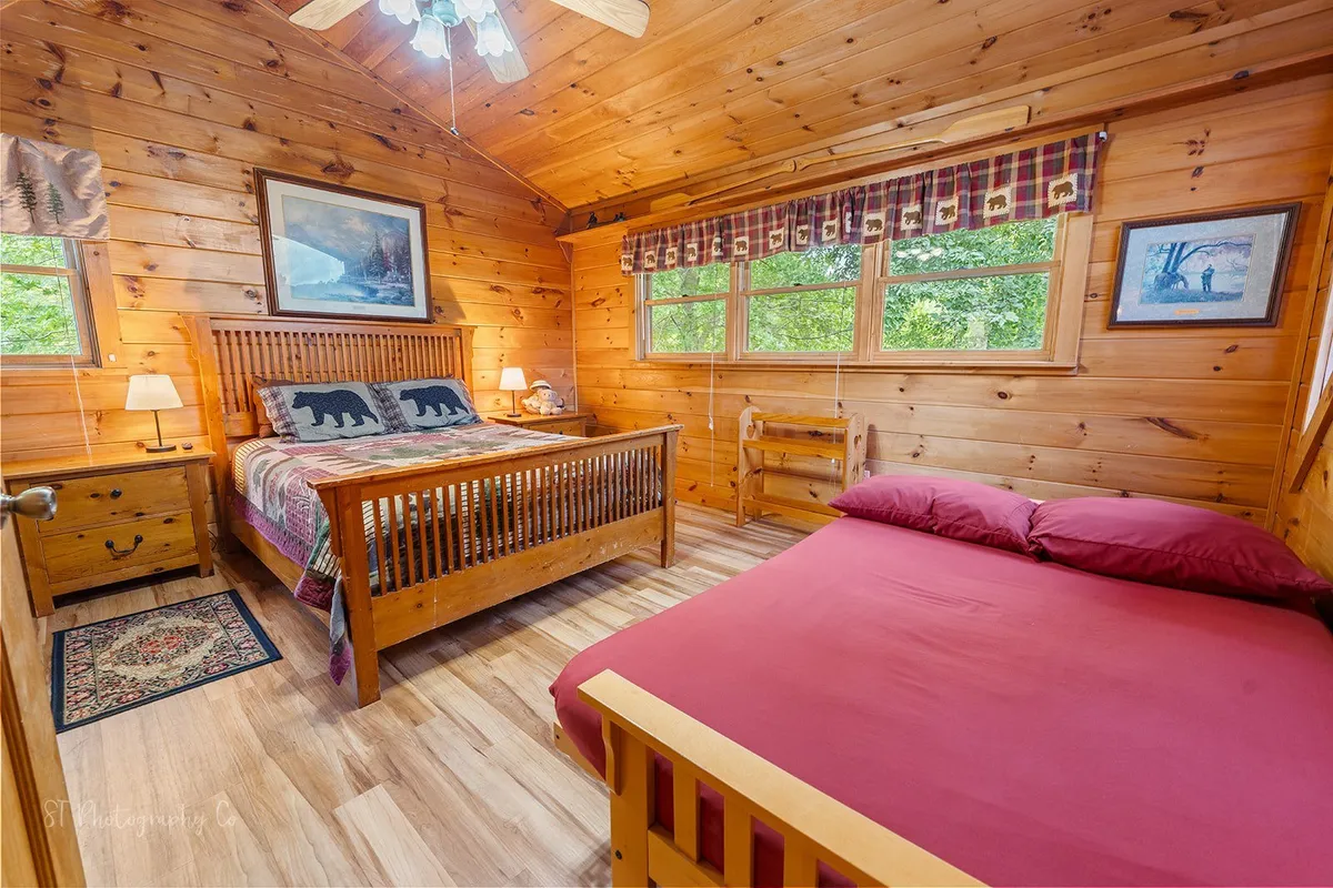 A warmly lit bedroom featuring a wooden bed frame adorned with bear-patterned bedding, complemented by wooden furniture and a framed artwork on the wall. Large windows offer views of the outdoors, creating a serene atmosphere.